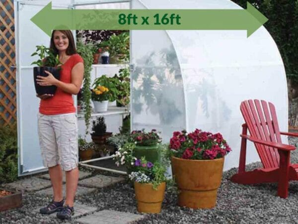 Solexx 8ft x 16ft Harvester Greenhouse G-416 - full view with plants and flowers in and out of the greenhouse - green arrow on top showing dimensions -  a woman outside carrying a pot with plants