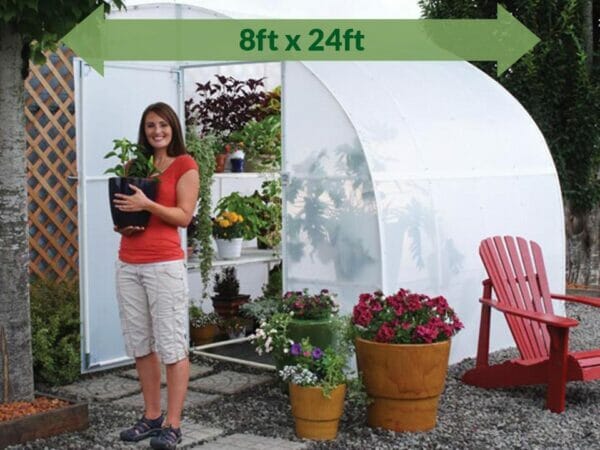 Solexx  8ft x 24ft Harvester Greenhouse G-424 - with plants - in a garden - a woman outside holding a pot with plant