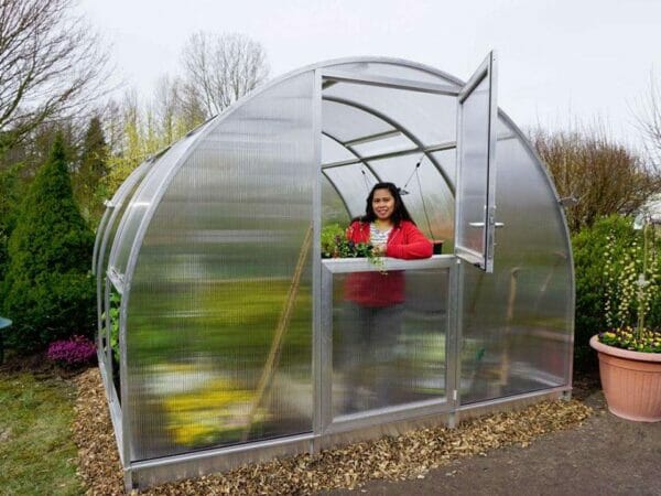 Front view of Arcus 3 Greenhouse - top section of door is open - a woman is standing inside