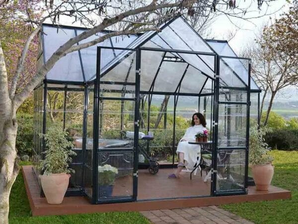 Palram Chalet 12ft x 10ft Hobby Greenhouse HG5400 - full view - open doors- in a garden - a woman sitting inside