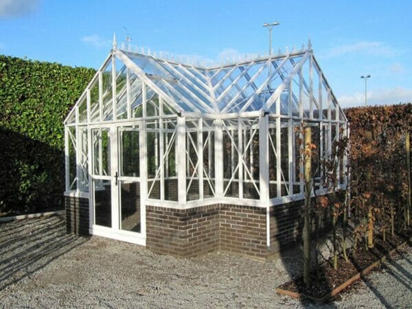 Janssens T-Shaped Royal Victorian Antique Orangerie, white frame with decorative roof ridge, on brick stem wall, in outdoor setting