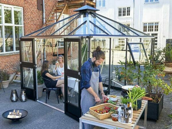 Juliana Oasis Greenhouse 10ft x 10ft Aluminum Outdoor setting. Two women inside the greenhouse and a man preparing meals outside