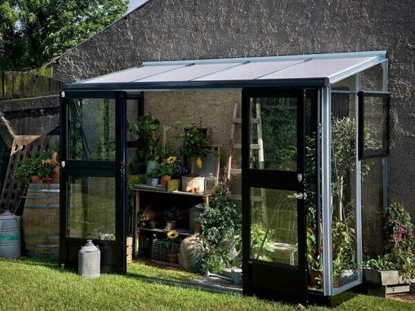 Juliana Veranda Lean-To Greenhouse 10ft x 5ft Aluminum 3mm toughened glass leaning against a wall. Open Doors. With Plants inside.