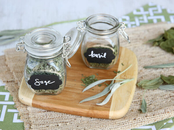 Glass jars filled with freeze-dried herbs on wooden cutting board, jars with blackboard labels for sage and basil