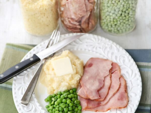Background: glass jars containing freeze dried foods, Foreground: White plate containing the re-hydrated foods (mashed potatoes, ham, and peas) with knife and fork