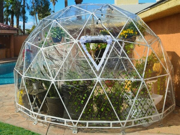 Rear view of geodesic dome with clear vinyl cover and steel frame, vent with optional bug screen, attachment hardware, plants and lighting on interior, pool in background