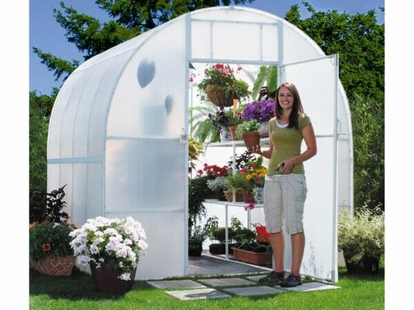 Woman standing at open door of arched greenhouse, interior shelves filled with flowering potted plants
