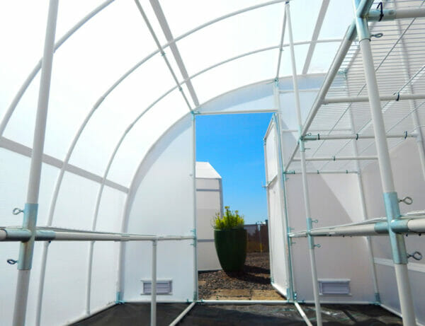Interior of lean-to greenhouse, facing door, door open, empty wire shelves lining both sides of greenhouse
