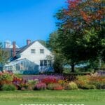 Image of flower garden in foreground and house and greenhouse in background, trees with fall color and the text: Autumn Tasks - How to Prepare Your Greenhouse for Winter