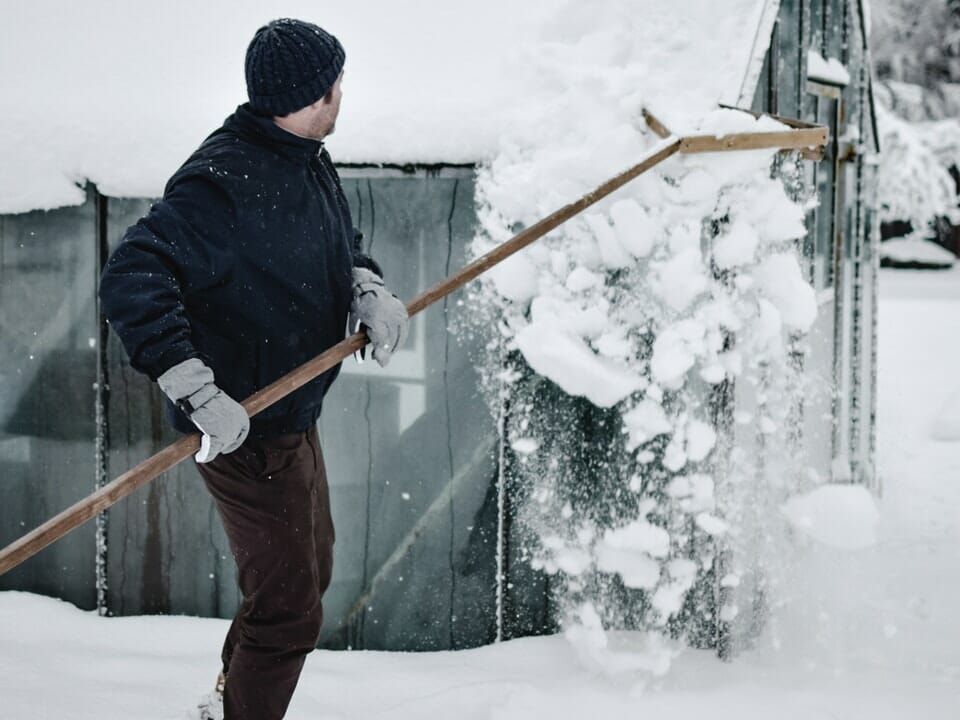 Man in jacket, hat, gloves with long handle snow removal tool, removing snow from greenhouse roof