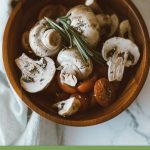 Wooden bowl on white tablecloth containing white mushrooms with tomatoes and rosemary herbs, with text: How to Grow Mushrooms in a Greenhouse