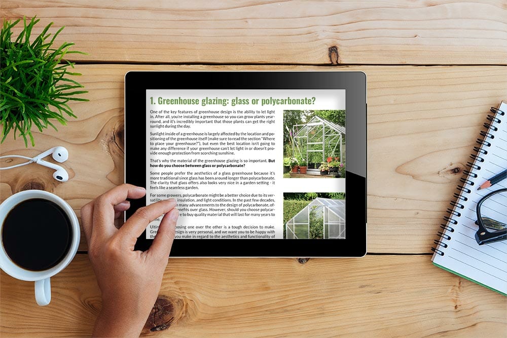 Tablet on a wooden table with plant, coffee, notebook, headphones, and hand zooming into the document which is a page of the eBook: Your Greenhouse, Your Choice - A Practical Guide To Picking Your Greenhouse Kit