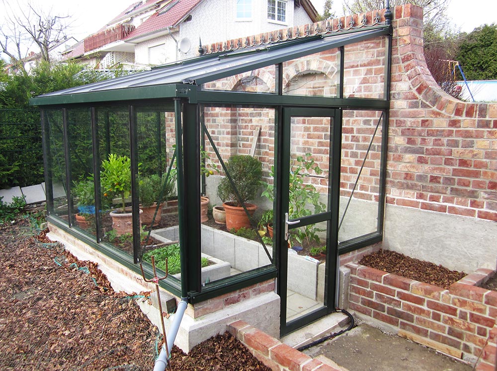 Green-framed Janssens Arcadia lean-to glass greenhouse on a stem wall installed on a brick wall