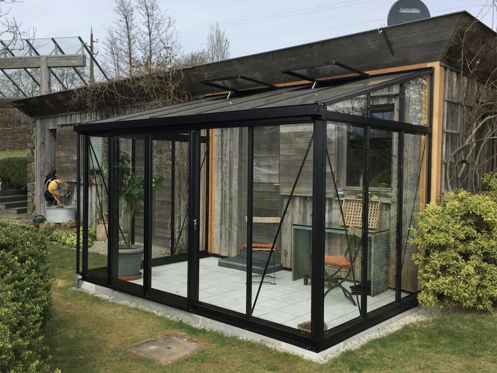 Black-framed lean-to glass greenhouse on a rustic house wall
