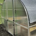 Exterior of a Riga polycarbonate greenhouse with an exterior shade and the Greenhouse Emporium logo at the top.