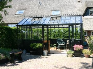 Front view of the Exaco Janssens VI34 Retro Victorian Greenhouse installed next to a house with a thatched roof