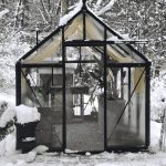 Exterior of a Janssens glass greenhouse covered in snow with the Greenhouse Emporium logo at the top.