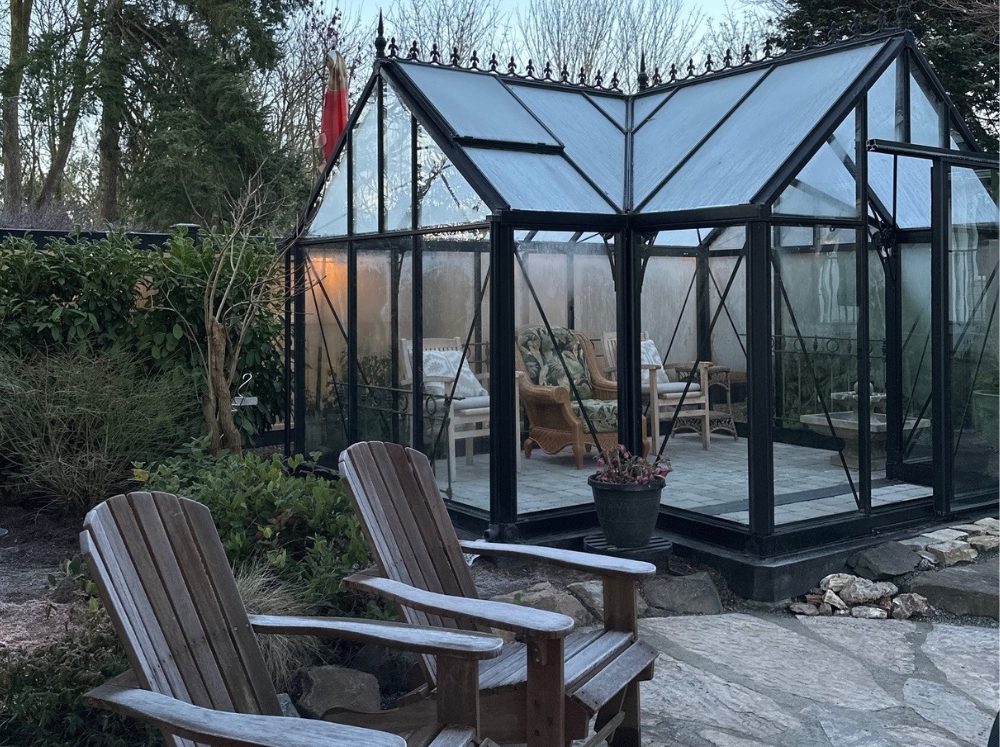 An antique glass greenhouse used as a lounging area with some lounge chairs; set in a small garden with two more chairs outside showing how to use a greenhouse as a lounge area