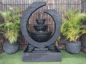Modern black hollow circular 45" tall eclipse fountain that is open at the top with two bowls sitting in the bottom of the circle. Water flows from the very top of the circle down to the other side of the top of the circle, down into the bowls and overflows into a 35” W x 28” D x 10” H rectangular base. Made of high-strength, durable glass-reinforced concrete, uses a 291 GPH pump, and weighs 531 lbs.