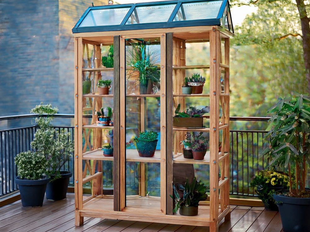 A small glass with wooden frame gable greenhouse on a back porch filled with potted plants. 