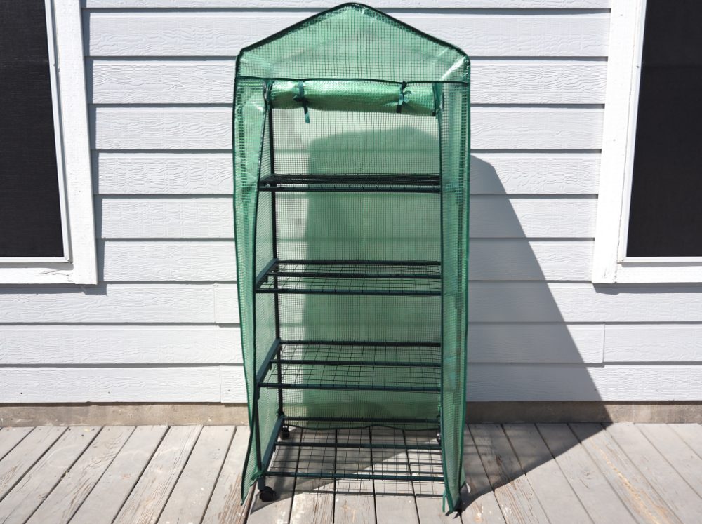 A small, but tall portable greenhouse on wheels with a plastic sheet covering it. It is placed on a home's deck.