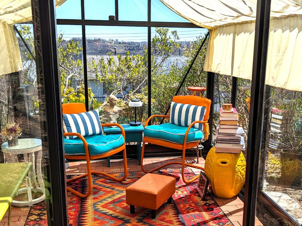 Greenhouse interior transformed into a cozy reading nook with a comfortable chair, side table, and books, bathed in natural light.