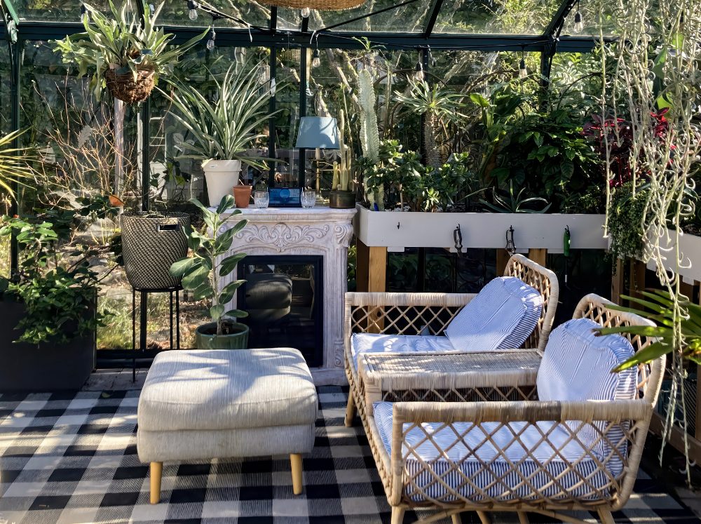 Opulent greenhouse lounge area with plush chairs, an ottoman, a cozy fireplace, and elegant shelving displaying potted plants.