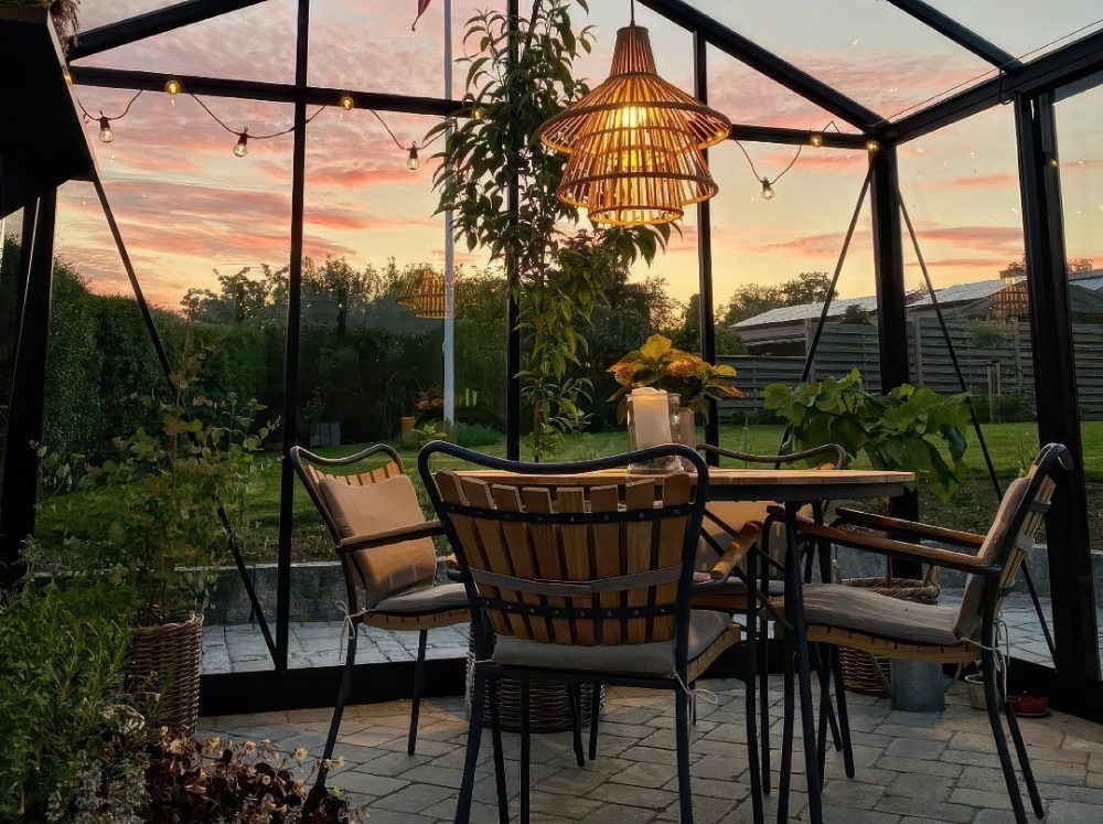 Greenhouse interior at night, illuminated by string lights and a table lamp, with comfortable seating for an inviting evening retreat.