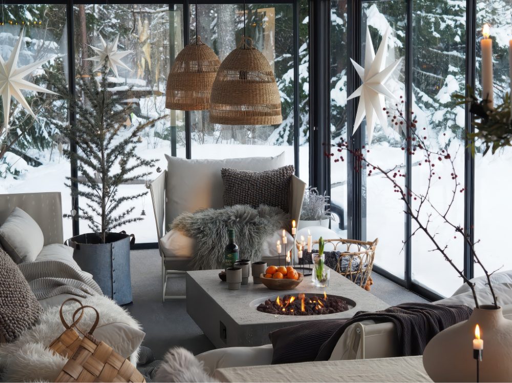 Greenhouse interior set up as a warm winter lounging area with plush sofas, throws, and subtle holiday decor, looking out onto a snowy landscape.
