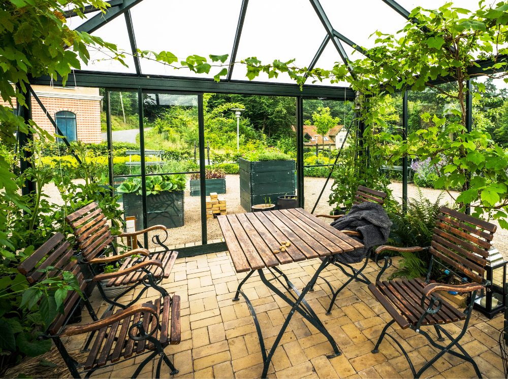 Spacious greenhouse interior featuring a rustic wooden dining table and chairs, surrounded by lush plants, set up as an inviting indoor picnic area.