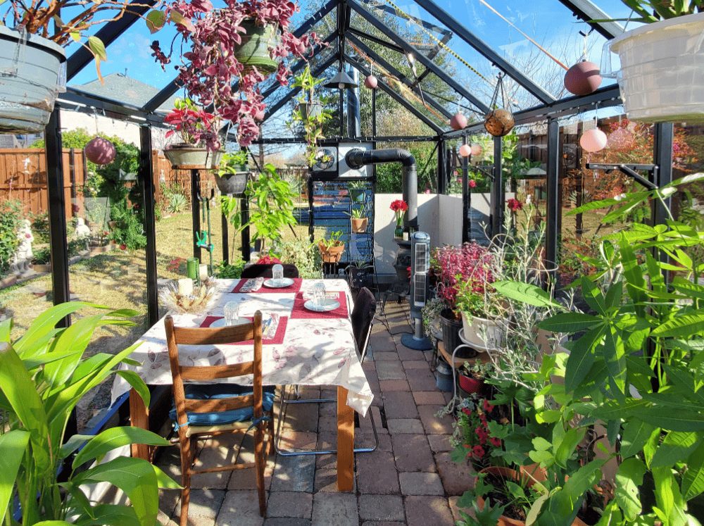 Greenhouse interior set up as a charming dining room with a table, chairs, and blooming plants, featuring a cozy wood stove nearby.