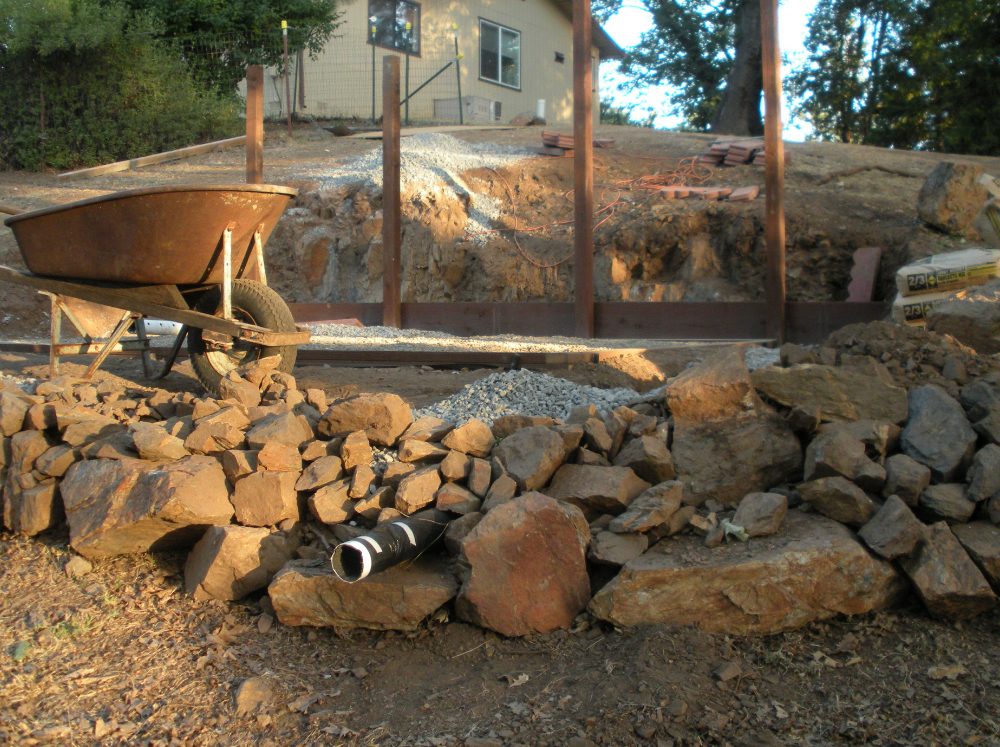 Construction site for a greenhouse foundation, showing excavated earth, a pile of large rocks, and wooden formwork for the base, with a wheelbarrow in the foreground.