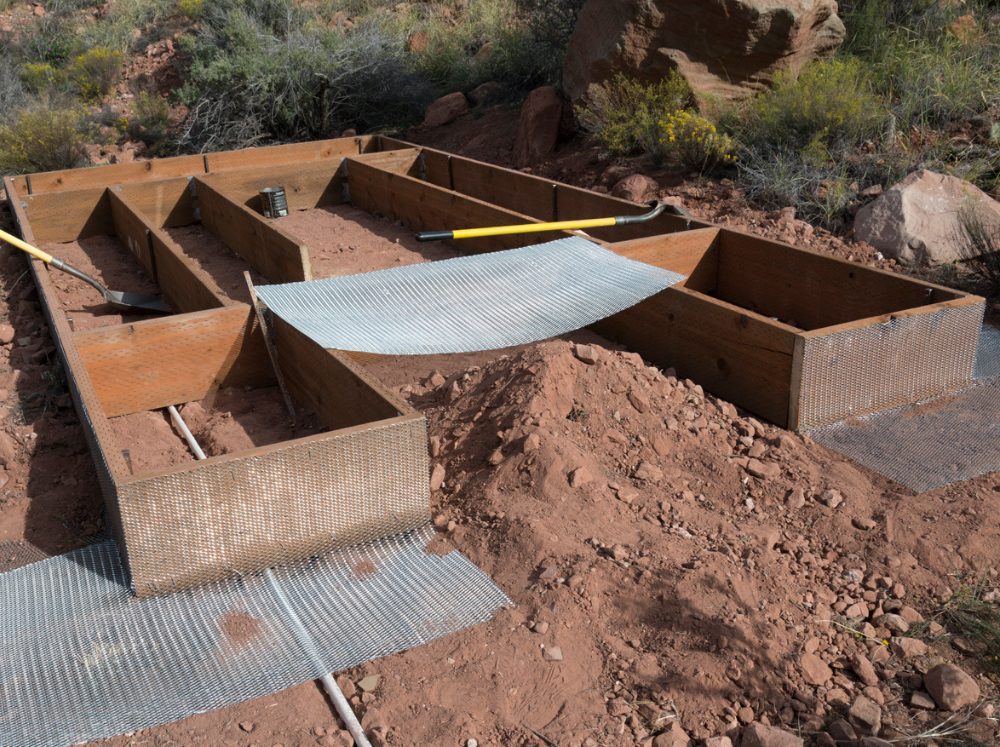 Greenhouse foundation under construction, showing a zigzag pattern of wooden raised beds lined with mesh, set on reddish soil in a desert-like landscape.