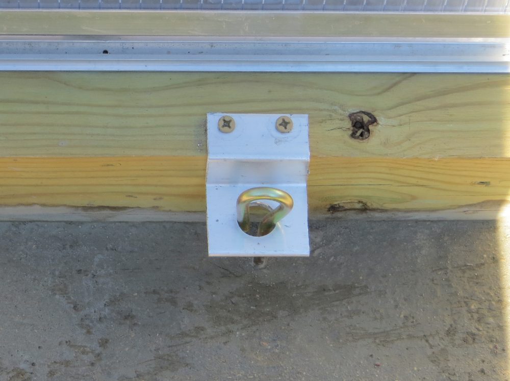 Close-up of a metal anchor bracket with a gold eye bolt secured to a wooden foundation beam, indicating how a greenhouse structure is fastened to its base.