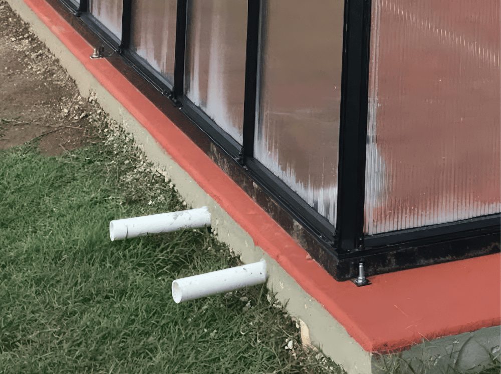 Close-up of a greenhouse base with a concrete foundation painted terracotta red, showing two white PVC pipes emerging from the base and resting on the grass.