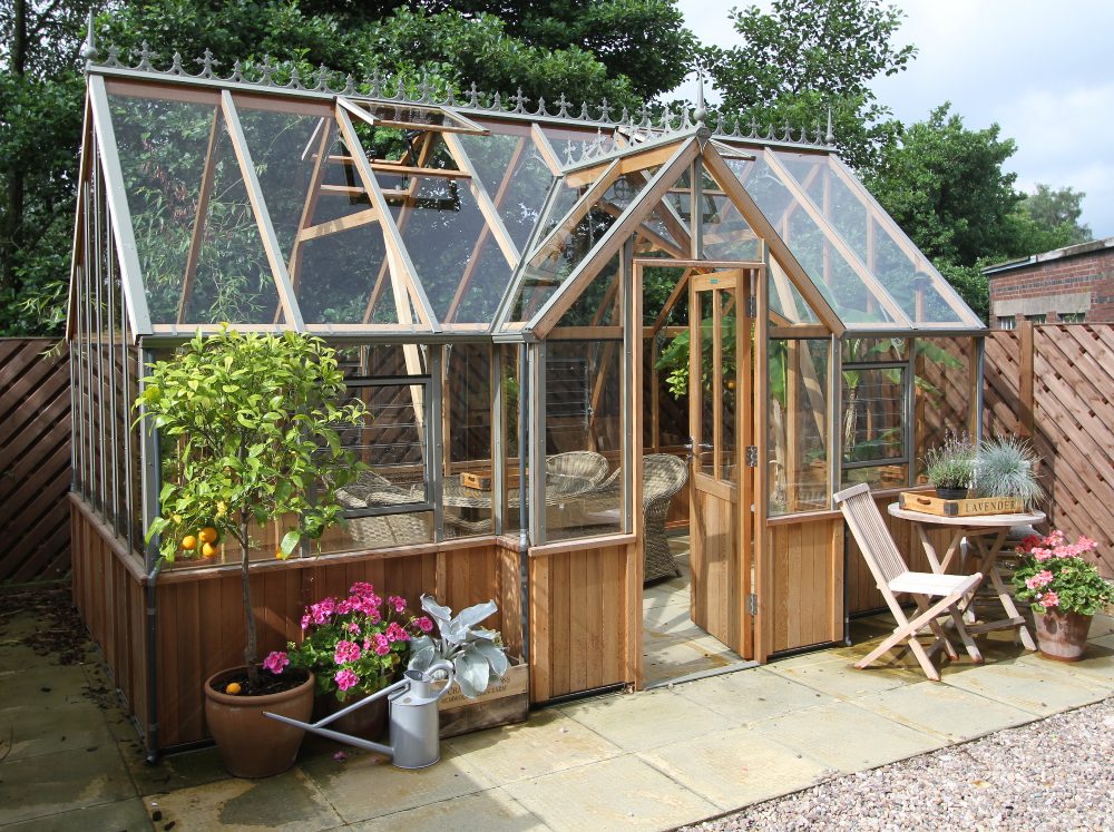 The best wooden greenhouse kit - Alton Cambridge greenhouse with glass panes set in a backyard with a cozy garden table and chairs in front.