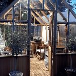 Front view of an Alton Cheltenham greenhouse in the evening with interior lighting and the greenhouse emporium logo at the top.
