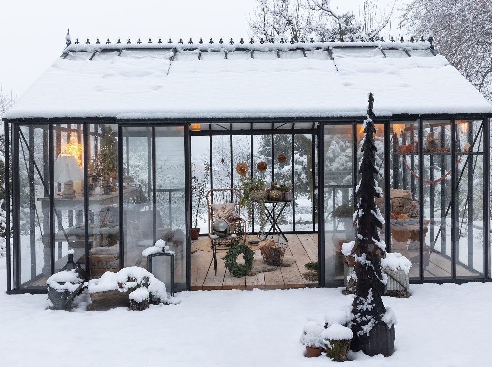 Glass gable greenhouse from the outside covered in snow with the doors open revealing holiday decorations on the inside.
