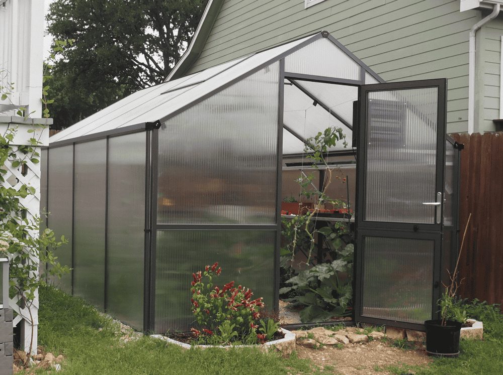 Front corner exterior view of a Nelke Pro polycarbonate greenhouse nestled between a fence and a home.