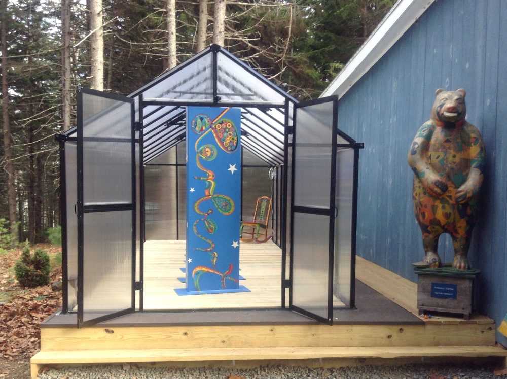 Tall thin paintings set up displayed inside of a greenhouse on a wooden deck and a bear statue next to it. 