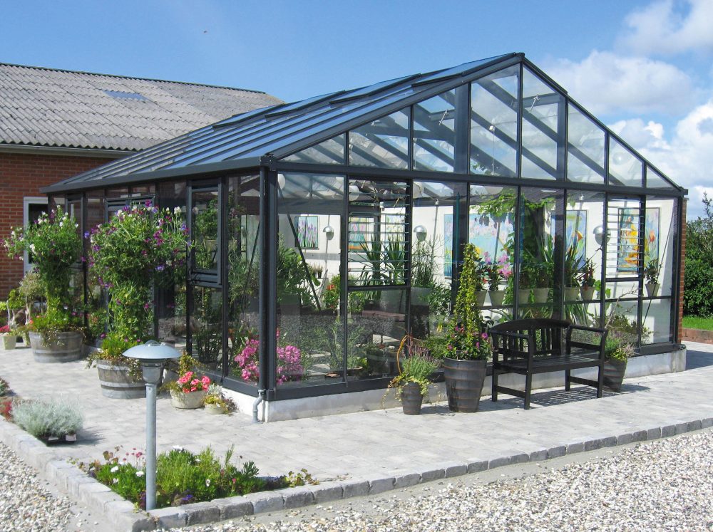 Front corner perspective of a glass Janssens Gigant greenhouse filled with and surrounded by plants.